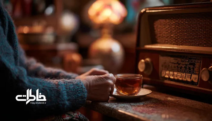 مادربزرگی نشسته در کنار رادیوی قدیمی با فنجان چای در دست، در فضایی گرم و نوستالژیک با نور زرد چراغ رومیزی؛ نمادی از آرامش خانه‌های ایرانی و خاطرات گذشته.